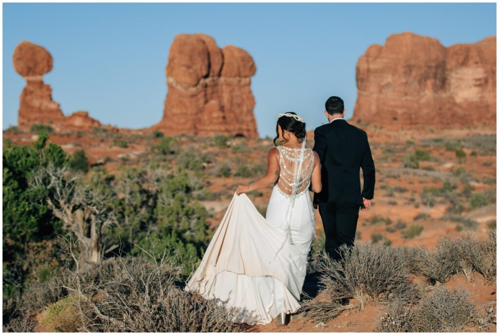 Bride and groom walk on a trail with Balanced Rock in the background.
