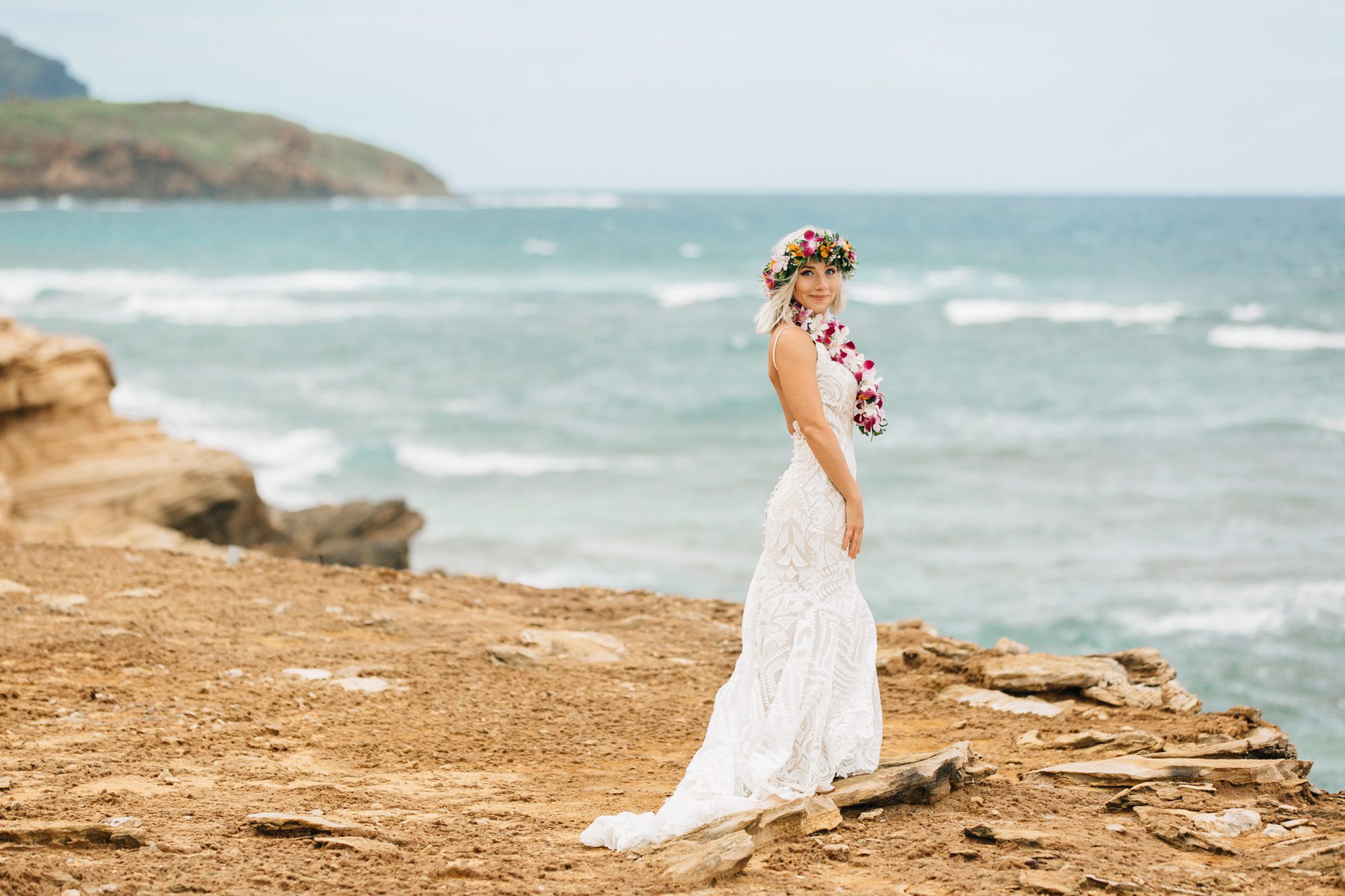 Bride has her photo taken on a cliff overlooking the ocean on Kauai. She wears a bright flower crown and lei.