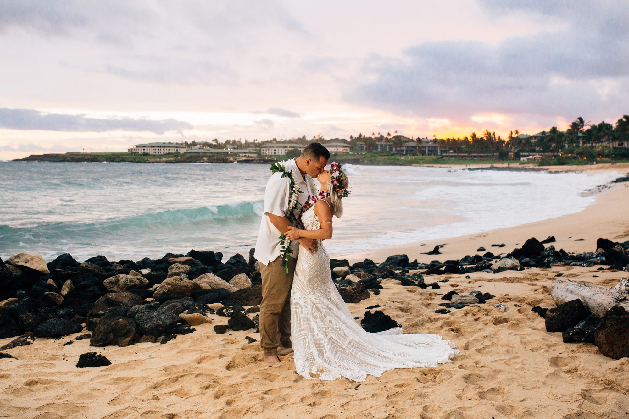 Bride and groom kiss on a rocky beach with the Hawaiian sunset in the background.
